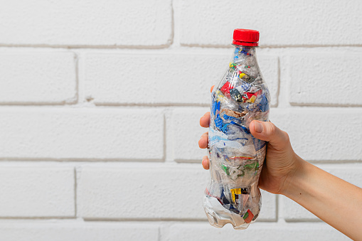 A hand holding a PET bottle filled with plastic waste for ecobricking with white brick wall on the background and space for text and copy
