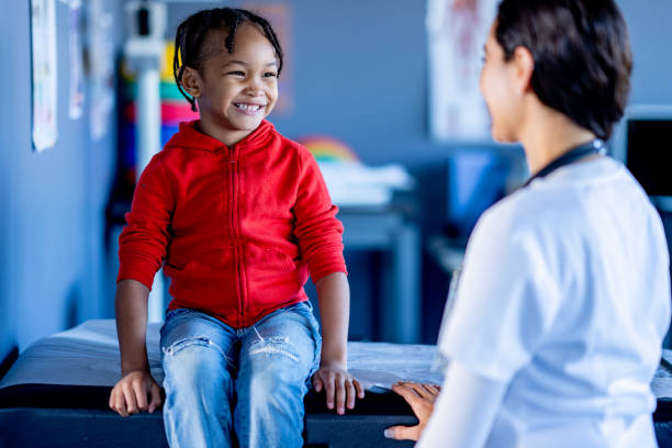 Pediatric Medical Appointment A sweet little boy of African decent, sits up on an exam table during a pediatric medical appointment. His female doctor is talking with him to put him at ease for the appointment. red medical scrubs stock pictures, royalty-free photos & images