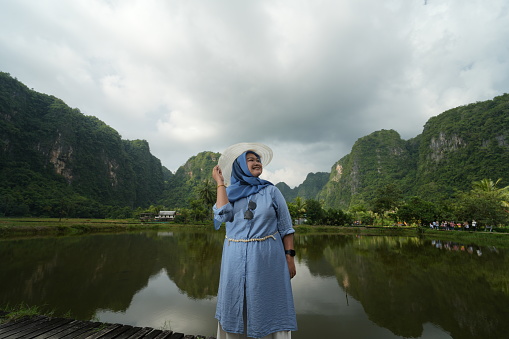 Asian model in hijab is posing with a hat in nature with mountains and water in the countryside
