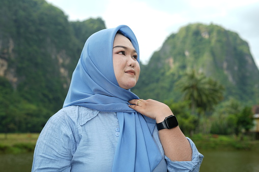 Asian model in hijab wearing blue shirt is posing in nature with mountains and water in the countryside as a backdrop