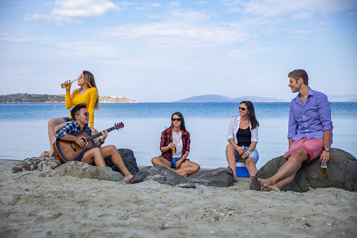 Group of happy young friends playing guitar, listening music, drinking beers on sandy beach party in sunny summer day against sea and cloudy blue sky. There are three girls and two boys sitting on rocks.