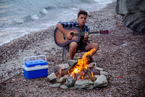 Young man playing guitar on the beach by the campfire.