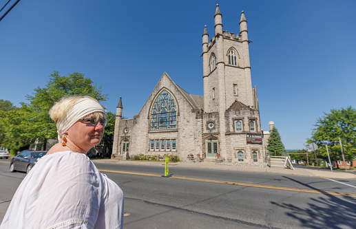 Mature Caucasian woman in a white blouse and a headscarf, standing on a street in Nazareth, Pennsylvania, with the historic St. Johns UCC Nazareth in the background. The church, known for its distinct architecture and cultural significance and heritage of the city