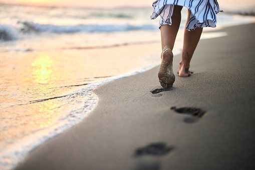 Close up of unrecognizable woman leaving footsteps in sand while walking on the beach at sunset. Copy space.