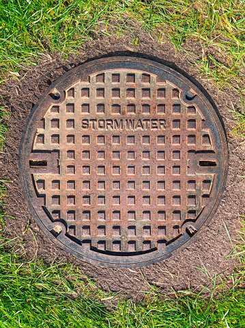 A rusty steel manhole cover for a stormwater drain, embedded in lush green turf in the summer, mitigating rainwater during extreme weather and storms.