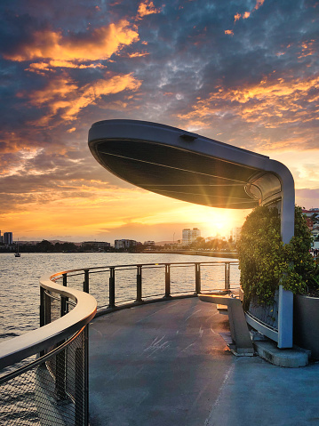 A stylishly designed shelter along a riverside pier with a beautiful sunset behind the city and river, illuminating a dramatic cloudscape in the sky.  The Lores Bonney Riverwalk along the Brisbane river in Brisbane, Australia.