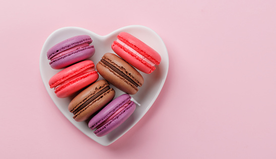 Various colorful macaroons in heart shaped bowl. Love sweets over pink background with copy space