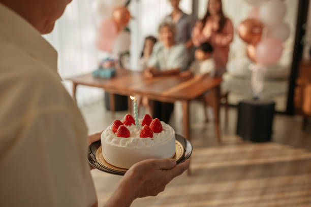 Senior man brings the birthday cake while the family is waiting gathered around the table.