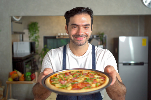 Cheerful male chef extends his arm to present a freshly baked pizza tray and graciously smiles at the camera positioned within the confines of the bustling kitchen.
