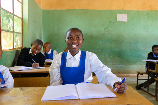 An African learner student sitting in a rural school classroom on a wooden desk keen to be educated.  Smiling Schoolgirl in uniform sitting in classroom looking at camera portrait.  Positive emotion.  Beautiful people. Malealea, Lesotho.