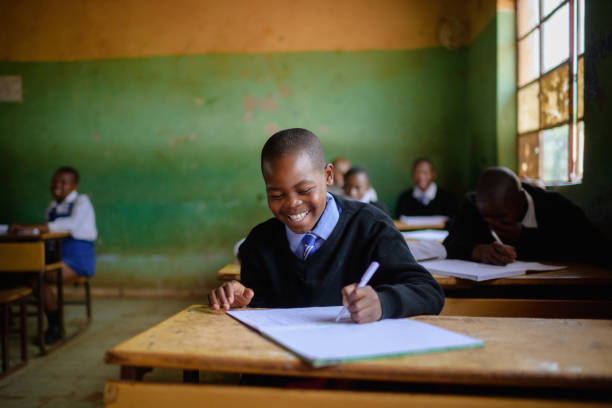 istock Cute Schoolboy in uniform sitting in classroom smiling whilst writing 2150086585