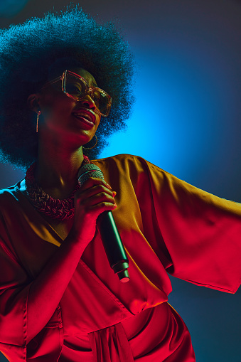 Soulful African-American singer in retro red attire and round glasses performs in neon light against gradient background. Concept of art, music, hobby, classical music and modern lifestyle. Ad