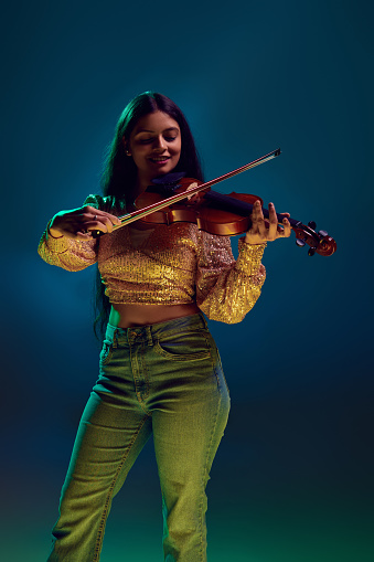 Artistic young Indian woman in glittery top enjoys playing violin in neon light against gradient background. Concept of art, music, hobby, classical music and modern lifestyle. Ad