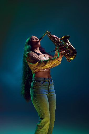 Side view photo of Indian woman, saxophonist performing solo in neon light against gradient background. Concept of art, music, hobby, classical music and modern lifestyle. Ad
