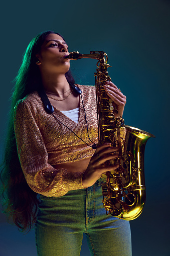 Intense Indian female musician with sax, wearing shiny apparel performing in neon light against gradient background. Concept of art, music, hobby, classical music and modern lifestyle. Ad
