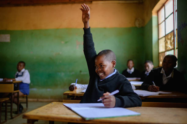 A Cute Male African learner student sitting in a rural school classroom on a wooden desk keen to be educated.  He has a pen in his hand while his other hand is raised and he is smiling.  Positive emotion.  Lifestyle Portrait. Beautiful people. Malealea, Lesotho.
