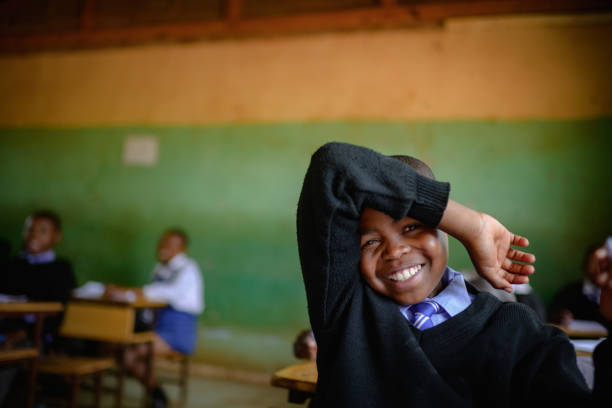 A Cute Male African learner student sitting in a rural school classroom on a wooden desk keen to be educated.  He has a pen in his hand while his other hand is raised and he is smiling shyly covering his eyes.  Positive emotion.  Lifestyle Portrait. Beautiful people. Malealea, Lesotho.