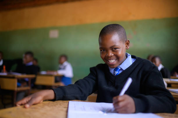 istock Cute Schoolboy in school uniform sitting in classroom looking at camera smiling and writing 2150087033