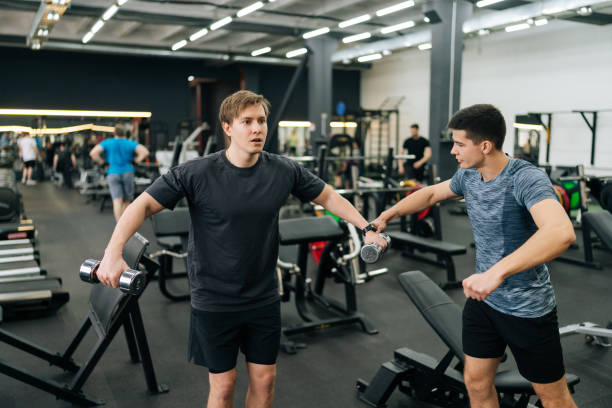 istock Tired beginner sportsman hard time doing lateral arm raise exercise with dumbbells with personal fitness trainer at gym. Sporty guy doing weight exercises 2150089213