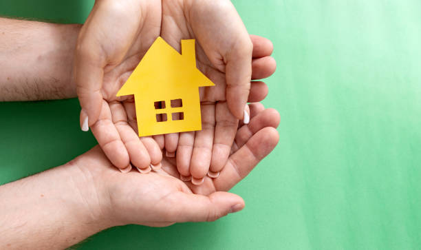 istock Two Hands Cradling a Paper House Cutout on Green Background 2150090471