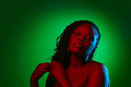 Elegant African woman with bare shoulders, well-kept spotless skin, posing on green studio background in neon light. Concept of natural beauty, ethnicity, self-care, wellness, positive emotions