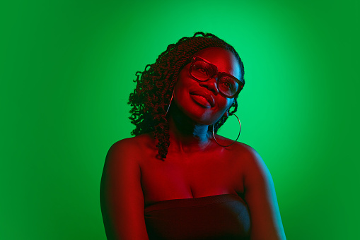 Attractive, elegant African woman in glasses, with curly hair and glowing spotless skin on green studio background in neon light. Concept of natural beauty, ethnicity, self-care, wellness, emotions