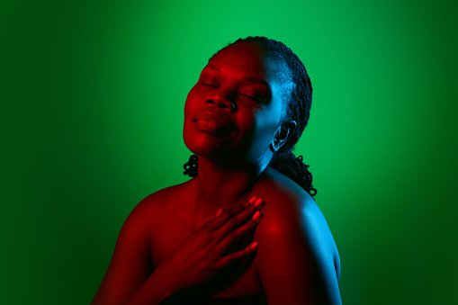 Elegant African woman with bare shoulders, well-kept spotless skin, posing on green studio background in neon light. Concept of natural beauty, ethnicity, self-care, wellness, positive emotions