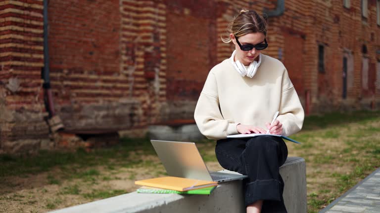 Woman looking at laptop screen while writing and spending time in campus yard