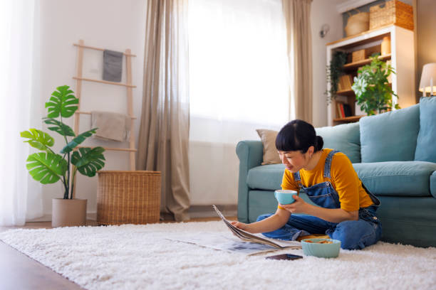 Beautiful mature Asian woman sitting on the living room floor, drinking coffee and reading newspapers while relaxing at home in the morning