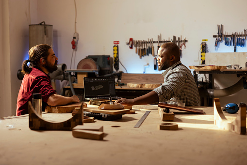 Woodworker and BIPOC coworker using CAD software on laptop to design wooden objects. Carpenter and african american man using program on notebook to plan furniture assembling in joinery