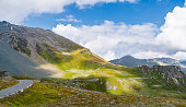 Panoramic View of Winding High Alpine Road Grossglockner Amidst Lush Green Hills, Rugged Mountains, and Blue Sky with Clouds
