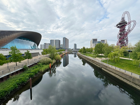 Canal in Queen Elizabeth Olympic Park, flowing past the London Aquatics Centra and ArcelorMittal Orbit, with residential tower blocks in the distance. April 2024.