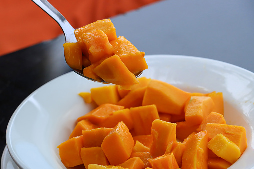 Stock photo showing close-up view of fruit with a metal spoon in white bowl with chopped mango.