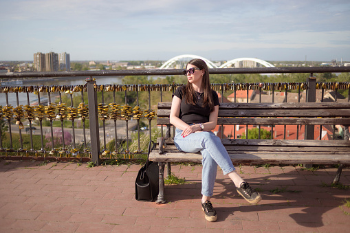 The woman is resting from the long walk in the city. She is sitting on the public bench, and enjoying the view of the city , and river.