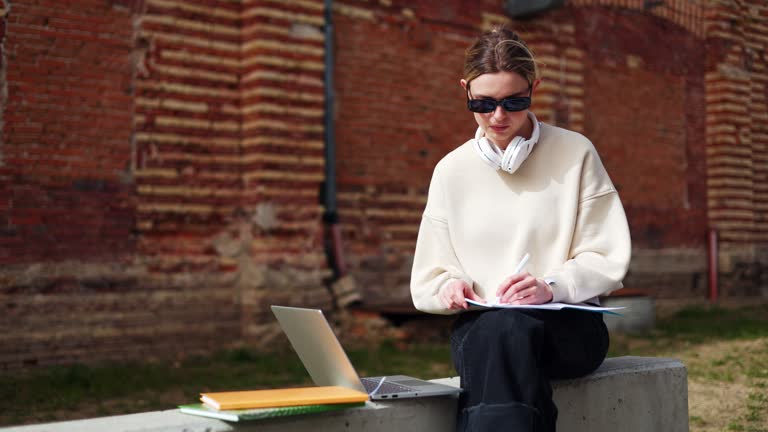 Female student in sunglasses making notes and using laptop while sitting outside