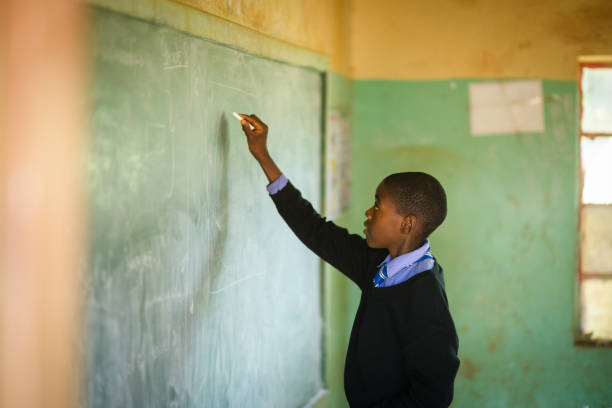 istock African Schoolboy Learner in school uniform writing with chalk on a chalkboard in a rural school classroom 2150104861