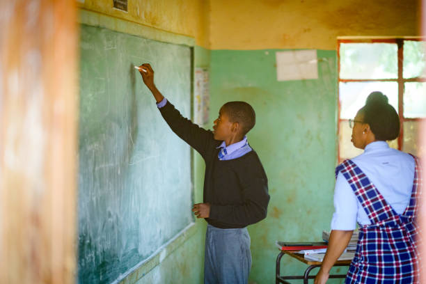 istock Schoolboy Learner in school uniform holding chalk with female teacher at chalkboard 2150104967