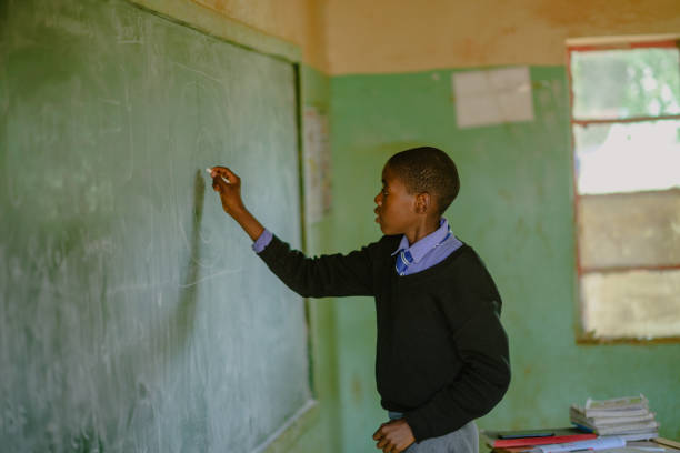 istock African Schoolboy Learner in school uniform holding chalk at chalkboard in a rural school classroom 2150105005