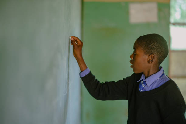 istock Copy Space African Schoolboy Learner in school uniform holding chalk at chalkboard in a rural school classroom 2150105047