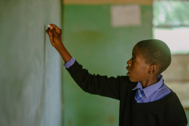 African Schoolboy Learner in school uniform writing on chalkboard in a rural school classroom