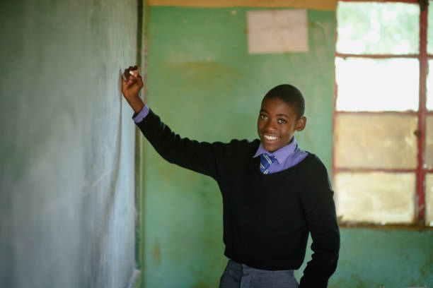 African Schoolboy Learner in school uniform holding chalk at chalkboard in a rural school classroom.  He is looking at the camera and is smiling.  Malealea Lesotho