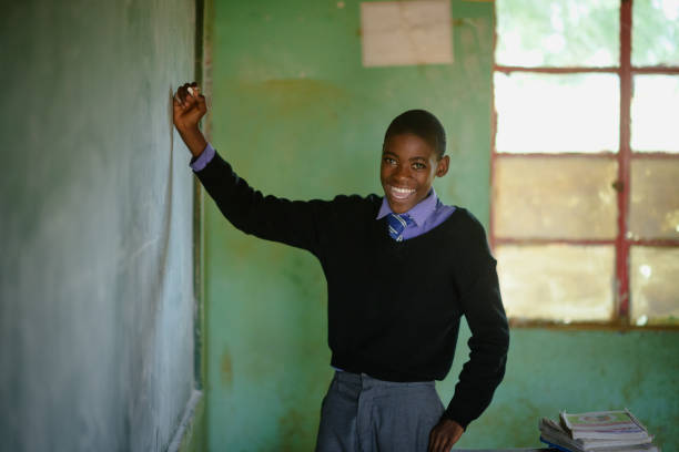 istock African Schoolboy Learner Portrait laughing in school uniform holding chalk at chalkboard in a rural school classroom 2150105461