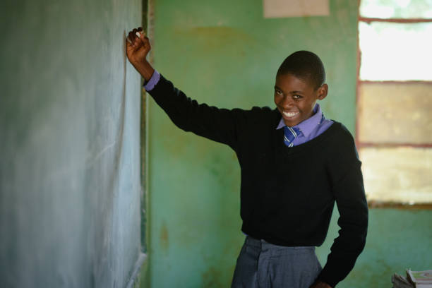 istock African Schoolboy Learner Portrait smiling shyly in school uniform holding chalk at chalkboard in a rural school classroom 2150105482