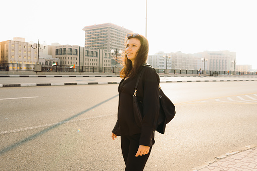 Female entrepreneur in black suit walking by the road looking for taxi with view of Arabian buildings during scenic sunset