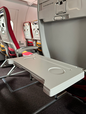 Stock photo showing an empty, airplane flip down table in row of seats in an economy class cabin of a passenger plane. A seat is viewed from the rear with a built-in fold down table.