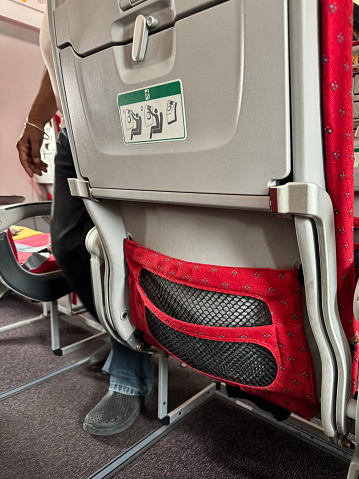 Stock photo showing close-up view of the interior of an airplane with rows of seats in an economy class cabin of a passenger plane. A seat is viewed from the rear with a built-in fold down table displaying an information label and an aircraft seat pocket net for documents and safety notices.