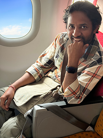 Stock photo showing close-up view of an Indian man sat in airplane cabin with a sunny blue sky and fluffy white clouds pictured through the plane window.
