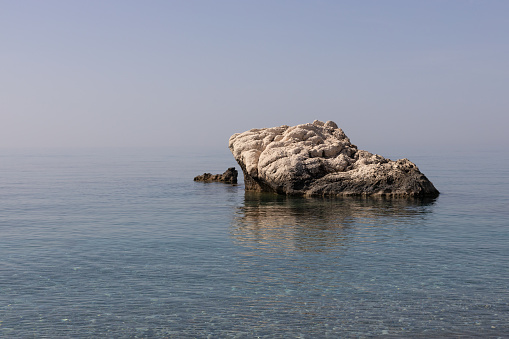 A weathered rock formation rises above the calm, clear blue waters of the Mediterranean Sea, under a soft sky.