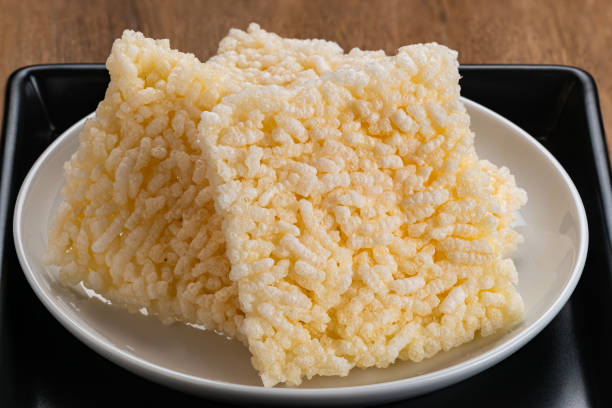 Closeup view pile of square shape crispy rice crackers in white ceramic dish placing in black ceramic tray on wooden table.