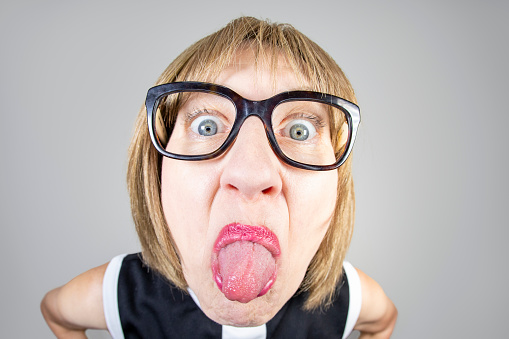 A fisheye image of a middle aged woman with short blond hair and black glasses sticking her tongue out in a childish way.